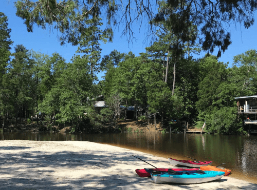 A serene river scene with colorful kayaks on a sandy shore, surrounded by lush green trees and clear blue skies.