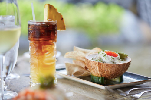 A tropical drink with a pineapple garnish beside a coconut bowl filled with seafood and garnished with herbs.