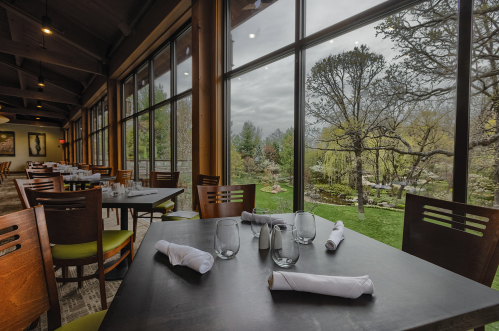 A dining area with tables set for guests, featuring large windows overlooking a lush garden and cloudy sky.
