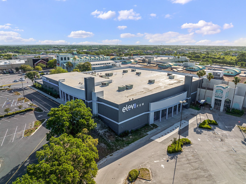 Aerial view of a large, vacant building with "elev8 fun" signage, surrounded by an empty parking lot and greenery.