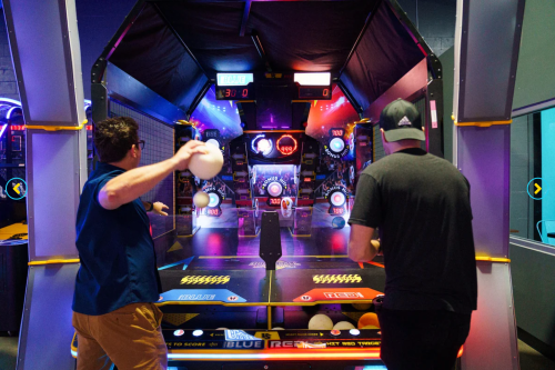 Two players compete in an arcade game, throwing balls at targets in a colorful, illuminated setting.