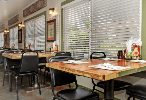 A cozy diner interior with wooden tables, black chairs, and bright windows covered by blinds.