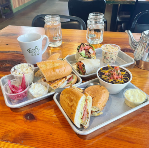 A wooden table with two trays of sandwiches, salads, and drinks, including a coffee cup and jars of water.