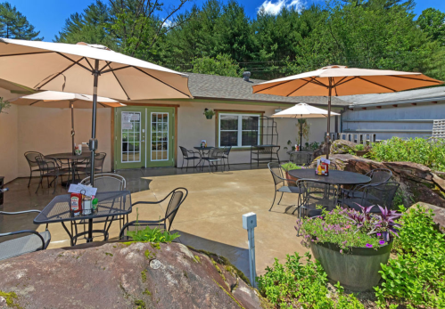 Outdoor patio with tables and umbrellas, surrounded by greenery and rocks, under a clear blue sky.