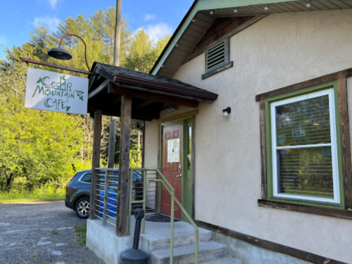 Exterior of Cedar Mountain Café, featuring a wooden sign, a door, and a car parked nearby, surrounded by greenery.