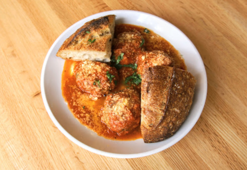 A bowl of meatballs in sauce, garnished with herbs, served with slices of crusty bread on a wooden table.