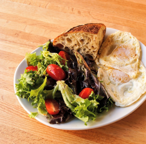 A plate with two fried eggs, mixed greens, cherry tomatoes, and a slice of toasted bread on a wooden table.