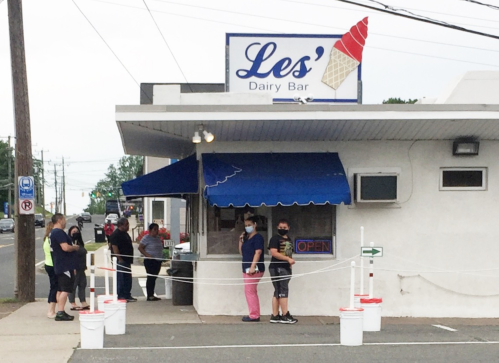 A small ice cream shop with a blue awning, featuring a line of customers waiting outside.