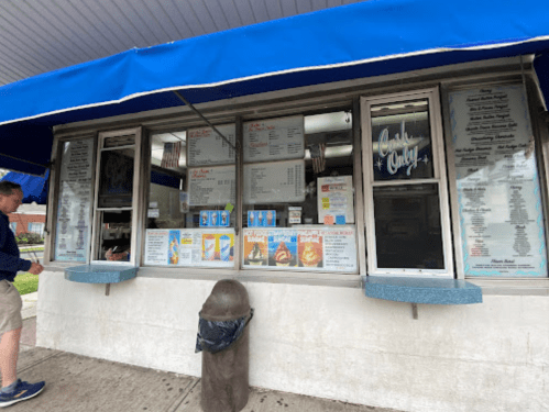 A small food stand with a blue awning, featuring a menu and a customer ordering at the window.