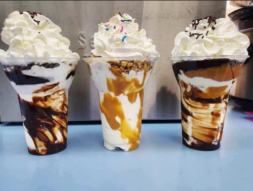 Three dessert cups with layers of whipped cream, chocolate, caramel, and sprinkles, served in clear plastic cups.
