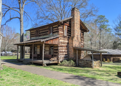 A rustic log cabin with a stone chimney, surrounded by trees and a grassy area on a sunny day.