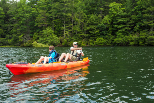 A man and a boy kayak on a calm lake surrounded by lush green trees.