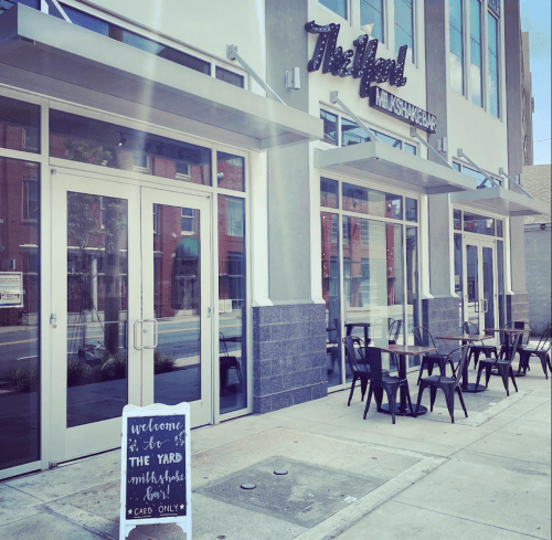 Exterior of a milkshake bar with a sign that says "Welcome to The Yard Milkshake Bar" and outdoor seating.