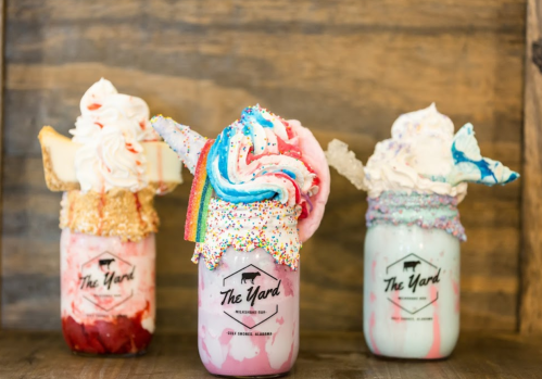 Three colorful milkshakes in jars topped with whipped cream, sprinkles, and candy, set against a wooden background.