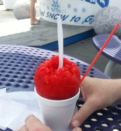A close-up of a red snow cone in a white cup, with a spoon and a straw, resting on a table.