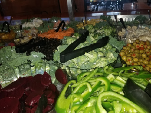 A colorful salad bar featuring various fresh vegetables, including greens, olives, and beets, with serving tongs.