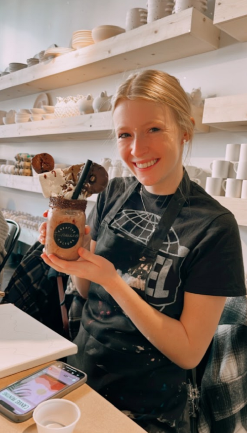 A smiling woman in an apron holds a large, decorated dessert drink with cookies and whipped cream in a cozy café setting.
