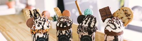 Four colorful milkshakes topped with whipped cream, cookies, and treats, displayed on a wooden counter.