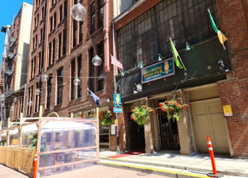 Entrance of a pub with hanging plants and flags, surrounded by brick buildings and outdoor seating.