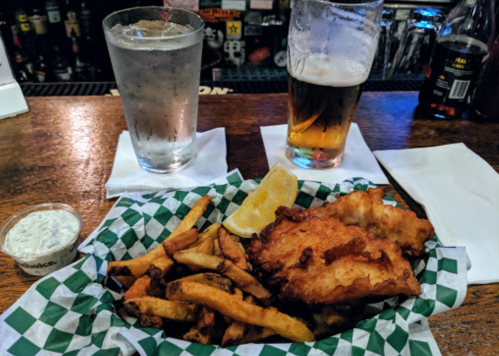 A basket of fried fish and fries with a lemon wedge, two glasses of water, and two beers on a bar counter.