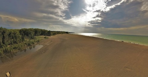 A wide view of a sandy beach with calm water, trees in the background, and a cloudy sky above.