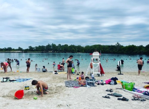 A busy beach scene with people swimming, lounging on towels, and a lifeguard chair near a calm water body under cloudy skies.