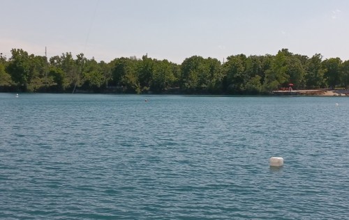 A calm blue lake surrounded by green trees under a clear sky, with a buoy floating in the water.