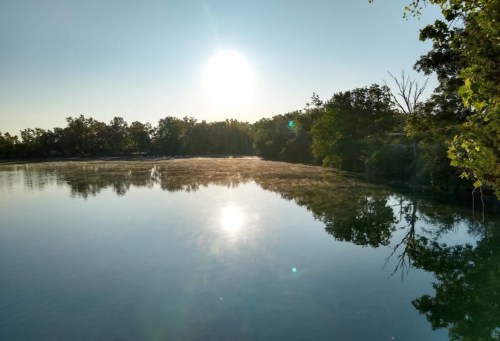 A serene lake at sunrise, with mist rising from the water and trees reflecting on the calm surface.