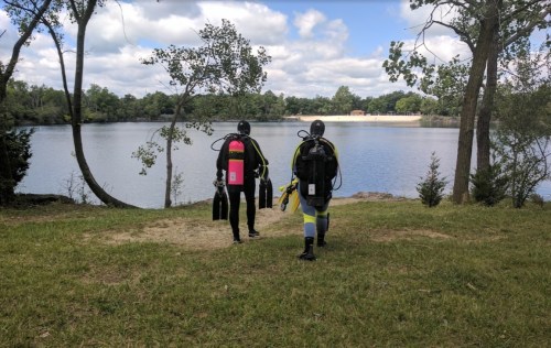 Two divers in wetsuits walk towards a lake, carrying scuba gear, with trees and clouds in the background.