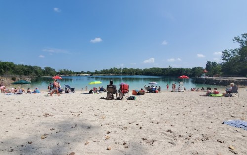 A sandy beach scene with people relaxing under umbrellas by a calm lake on a sunny day.