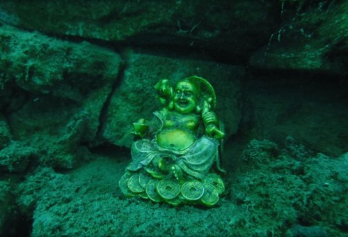 A small golden statue of Buddha submerged in green-tinted water, surrounded by sand and rocks.