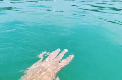 A hand reaching out into clear turquoise water, creating gentle ripples on the surface.