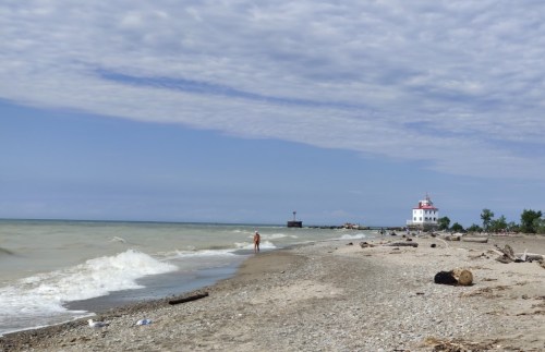 A sandy beach with gentle waves, a distant lighthouse, and a cloudy sky. A person walks along the shore.