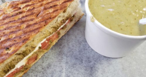 A toasted sandwich with tomatoes and cheese next to a cup of green soup on a gray surface.
