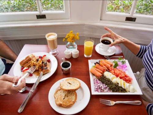 A table set with a breakfast spread: waffles, smoked salmon, bagels, drinks, and flowers in a cozy setting.