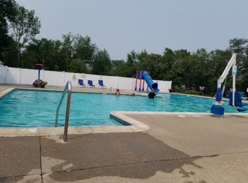 A swimming pool with a slide, surrounded by trees, and a few people enjoying the water on a sunny day.