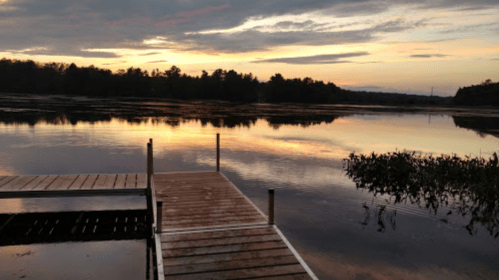 A serene lake at sunset, with a wooden dock extending into calm waters and trees silhouetted against the colorful sky.