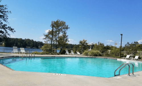 A clear blue swimming pool surrounded by lounge chairs, with a lake and trees in the background under a sunny sky.