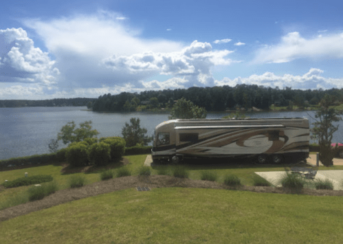 A luxury RV parked by a serene lake, surrounded by greenery and under a partly cloudy sky.