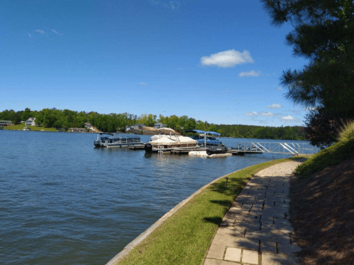 A serene lake scene with boats docked, surrounded by green trees and a clear blue sky. A pathway runs along the water's edge.