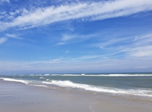 A serene beach scene with gentle waves, a clear blue sky, and wispy clouds reflecting a peaceful coastal atmosphere.