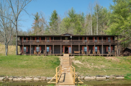 A rustic wooden lodge with multiple balconies, surrounded by trees and a small pathway leading to a waterway.