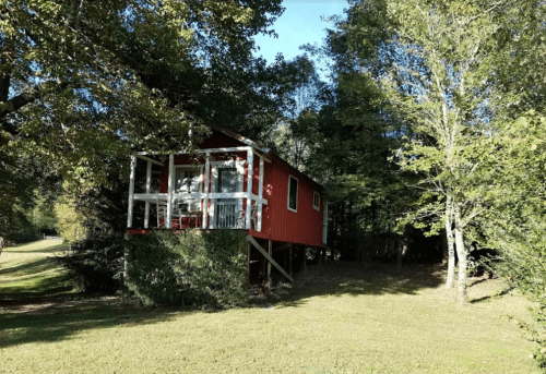A red cabin on stilts surrounded by trees and grass under a clear blue sky.