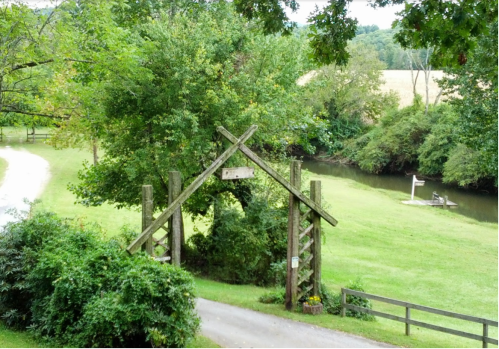 A rustic wooden archway leads to a grassy area by a calm stream, surrounded by trees and fields.