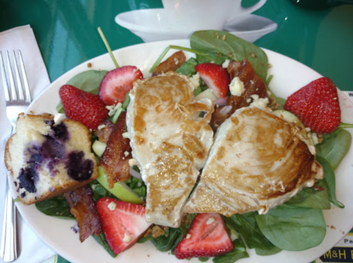 A plate of spinach salad topped with grilled chicken, strawberries, bacon, and a slice of blueberry bread on the side.