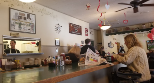 A cozy diner scene with patrons at the counter, a server in the kitchen, and Valentine's decorations hanging.