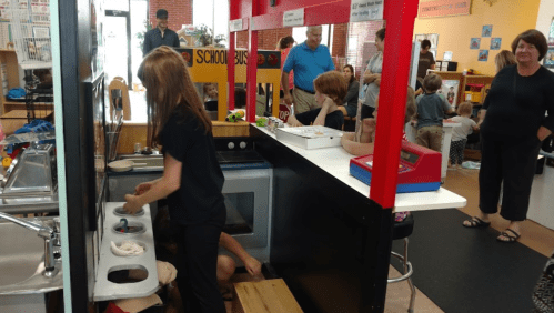 Children play in a classroom kitchen setup, engaging in imaginative play while adults observe nearby.