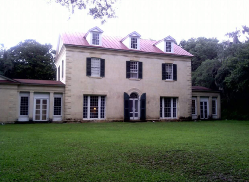 Historic two-story house with a red metal roof, white shutters, and large windows, surrounded by green grass and trees.