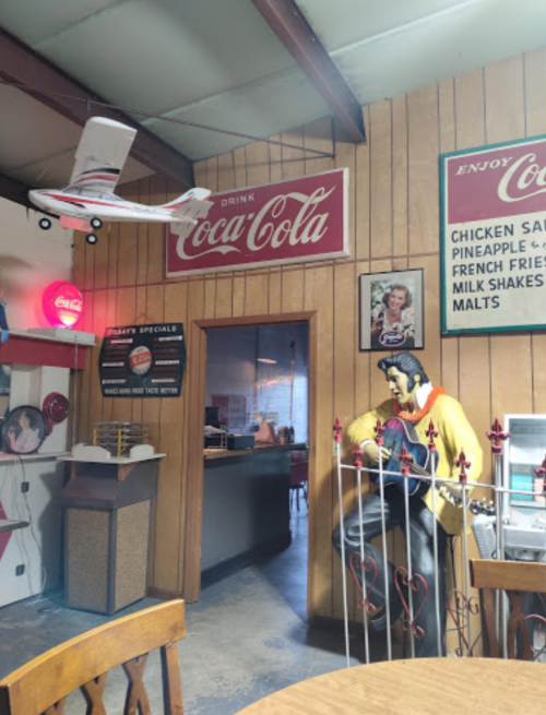 A retro diner interior featuring Coca-Cola signs, a model airplane, and a statue of Elvis near a table.