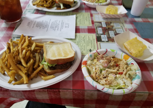 A plate with a sandwich, fries, and a colorful salad, with a dessert and drinks in the background on a checkered tablecloth.